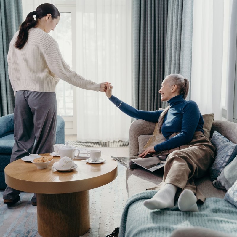 Woman holding the hand of an older woman reclining on a sofa beside a tea table