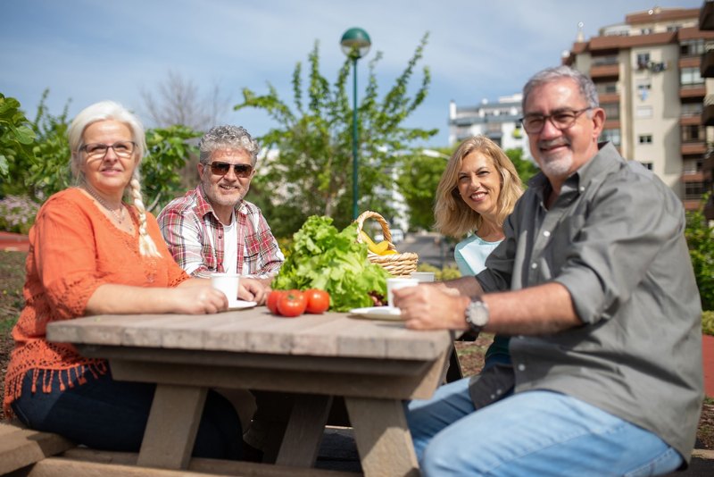 Four older adults smiling around a wooden picnic table with salad and tomatoes in a sunny park