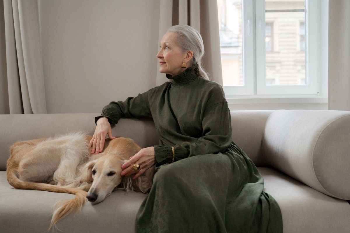 Older woman in a green dress on a beige sofa beside a long-haired dog lying curled up
