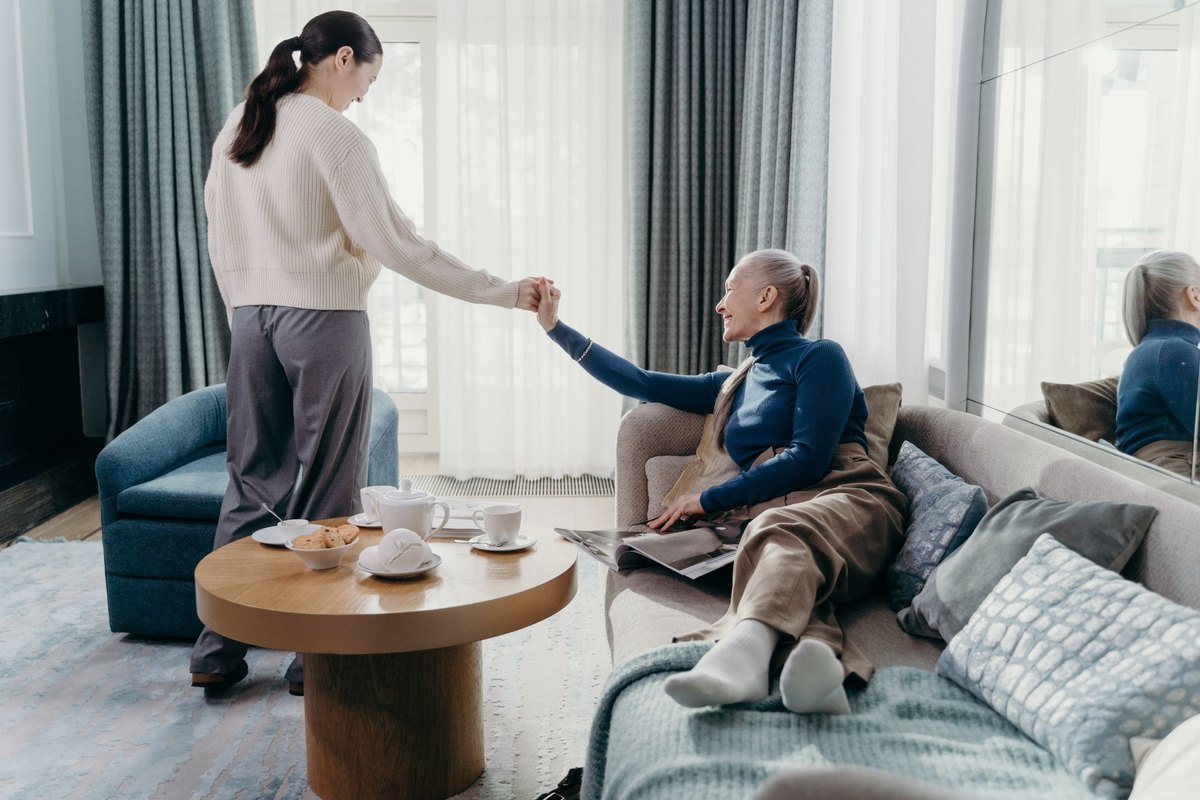 Woman in a cream sweater holding hands with an older woman resting on a sofa beside a tea table