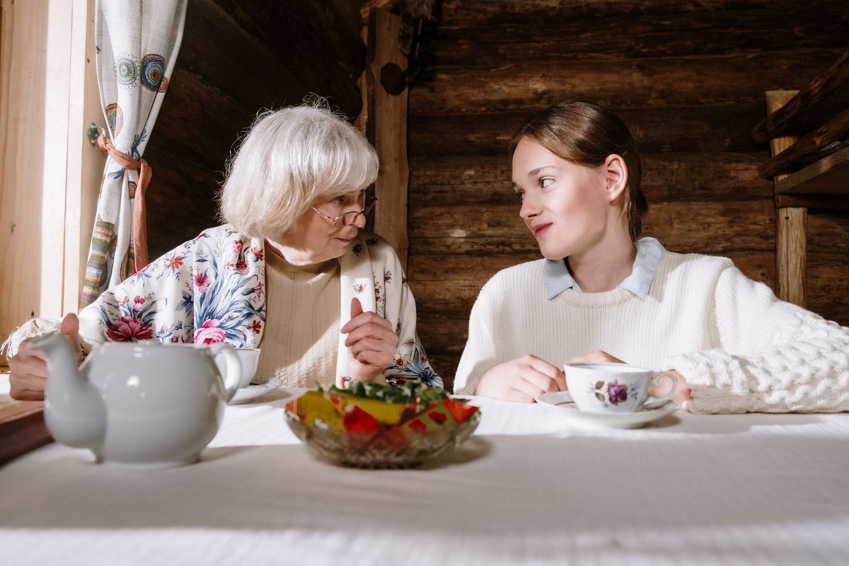 Older woman and younger woman sharing tea at a table with a teapot and fruit bowl