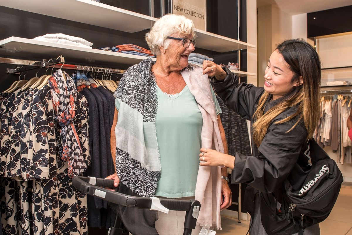 Older woman in a scarf holding a walker while a younger woman adjusts her scarf in a clothing store