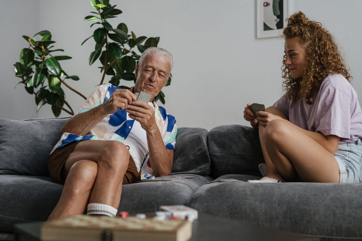 Older man holding playing cards on a gray sofa beside a young woman who holds cards at the other end