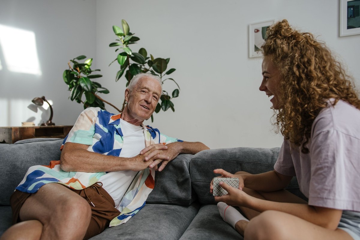 Older man smiling on a gray sofa while a young woman laughs beside him
