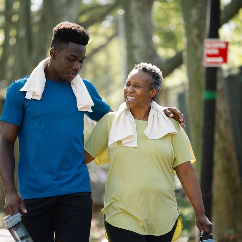 Older woman and younger man walking arm in arm with towels around their necks on a park path